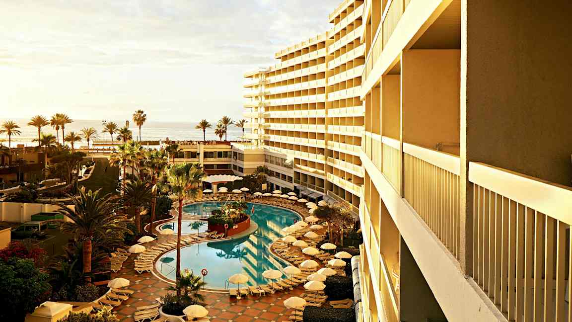 En utsikt over et hotellbasseng omgitt av solsenger og parasoller, med palmetrær ved siden og havet i bakgrunnen. (AI-generert tekst)