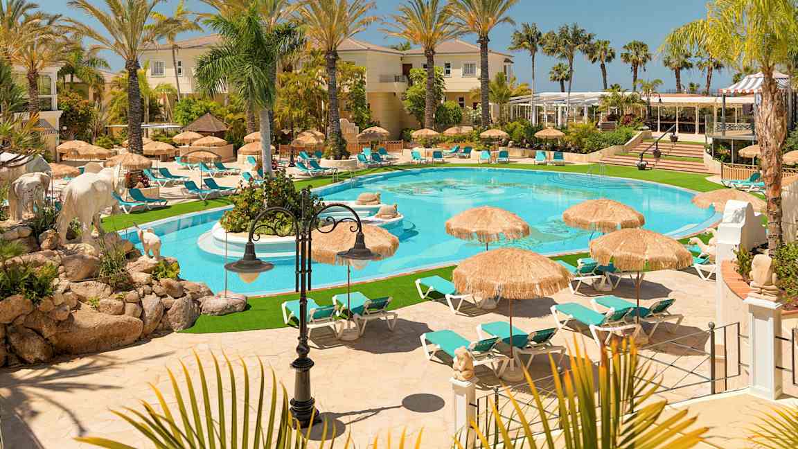 Et bilde av et luksuriøst feriested med et basseng, omgitt av solsenger og parasoller, omringet av palmer og bygninger. (AI-generert tekst)