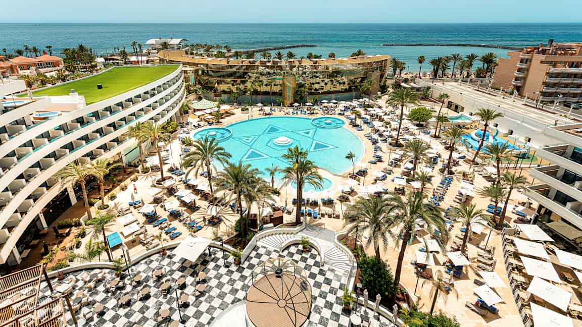 En utsikt over et stort hotellbasseng omgitt av solsenger, parasoller, palmer og bygninger med havet i bakgrunnen. (AI-generert tekst)