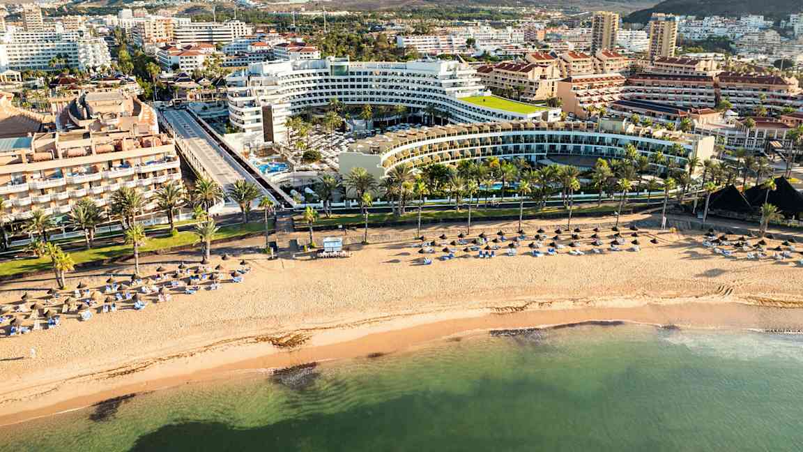 Bilde av en strand med parasoller og solsenger, omkranset av hotellbygninger og fjell i bakgrunnen. (AI-generert tekst)