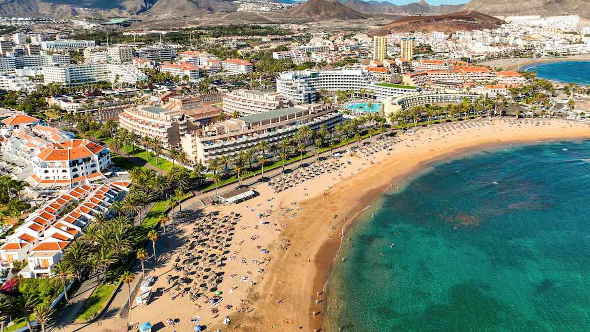 En sandstrand med solsenger og parasoller, omgitt av palmer og hotellbygninger i et turistområde. (AI-generert tekst)