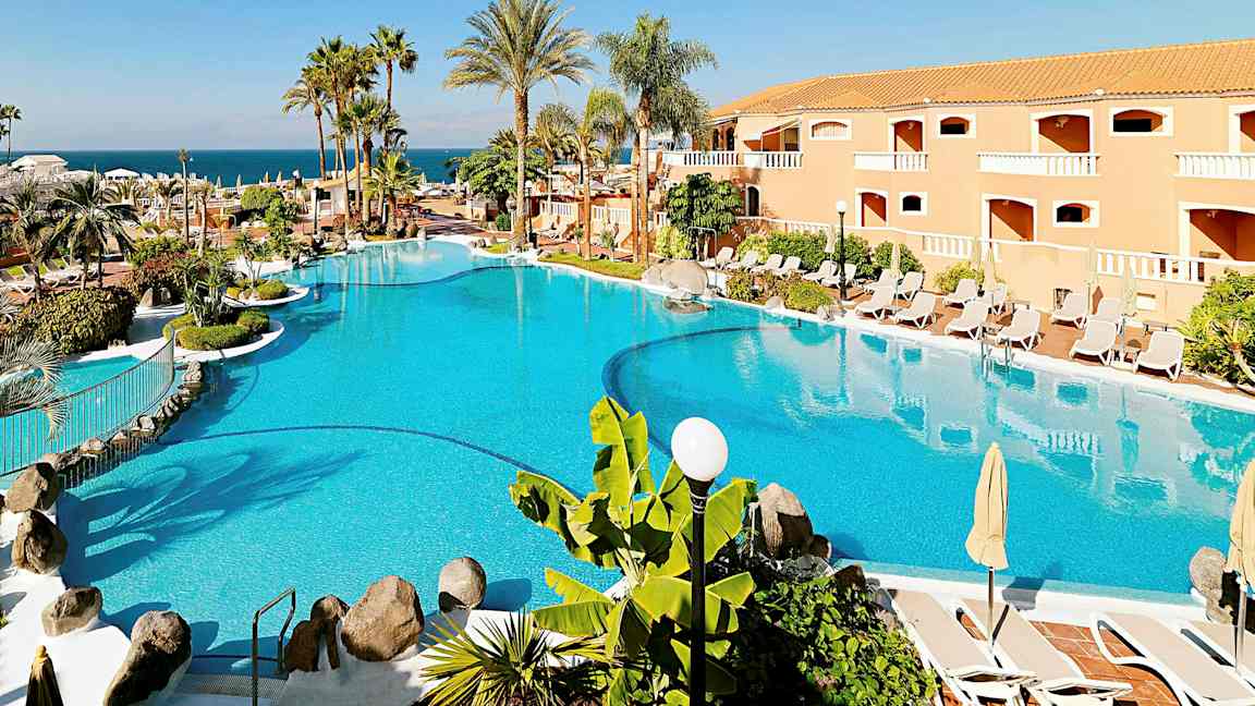 Et stort basseng omgitt av parasoller, solsenger, palmer og en bygning i bakgrunnen. (AI-generert tekst)