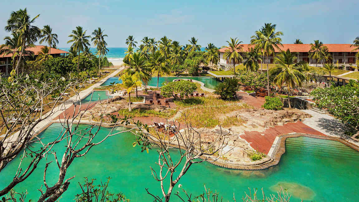 Hotellkompleks med bassenger, palmer og en sandstrand i bakgrunnen. (AI-generert tekst)