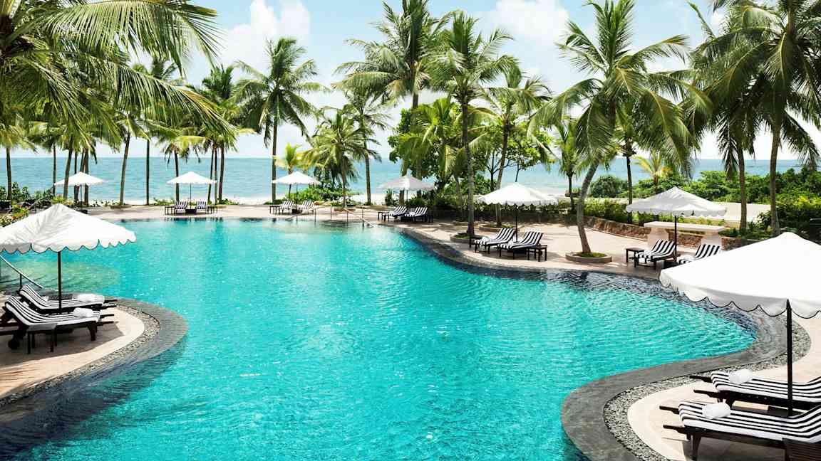 Bilde av et svømmebasseng omgitt av palmer og solsenger med parasoller ved havet. (AI-generert tekst)