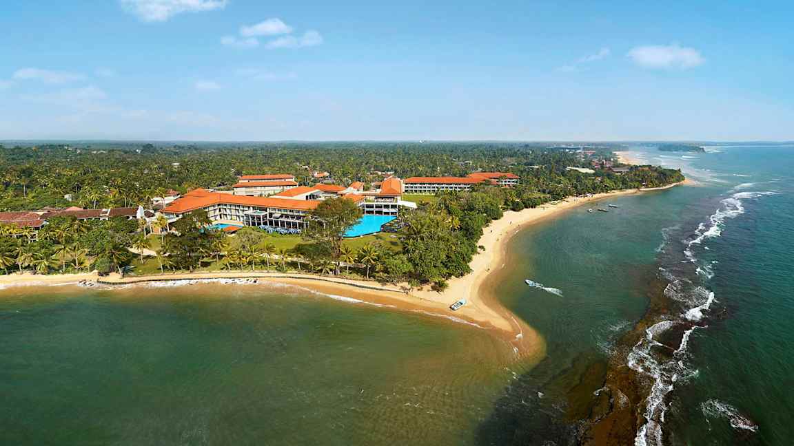 Et flyfoto av en kystlinje med sandstrand, hav og bygninger omgitt av vegetasjon. (AI-generert tekst)