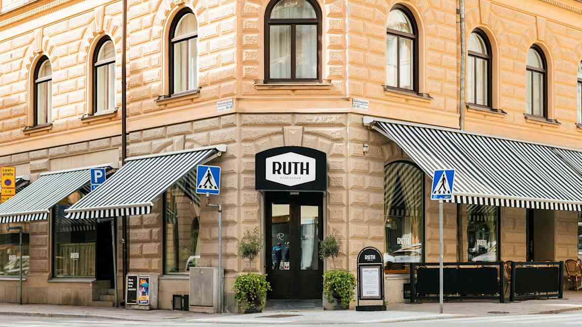 Et hjørnehus i klassisk stil med butikkens skilt Ruth over inngangen. Utstillingsvinduer med stripete markiser, gangvei og stolpeskilt er synlige i forgrunnen. (AI-generert tekst)