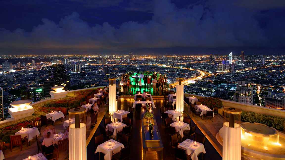 En takterrasserestaurant om kvelden med opplyste bord og utsikt over en by med lysende lys. (AI-generert tekst)