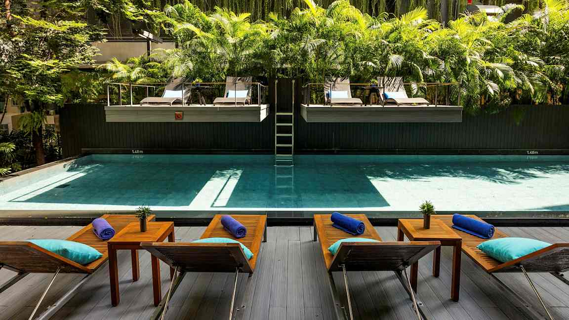 Et basseng med solsenger og bord foran, omgitt av grøntområder og palmer. (AI-generert tekst)
