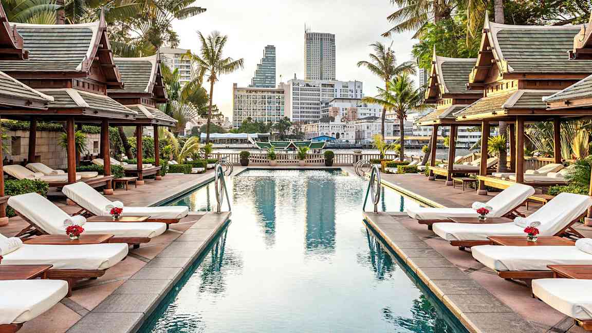 Et bassengområde omgitt av solsenger, palmer og paviljonger i tradisjonell stil med en by i bakgrunnen. (AI-generert tekst)