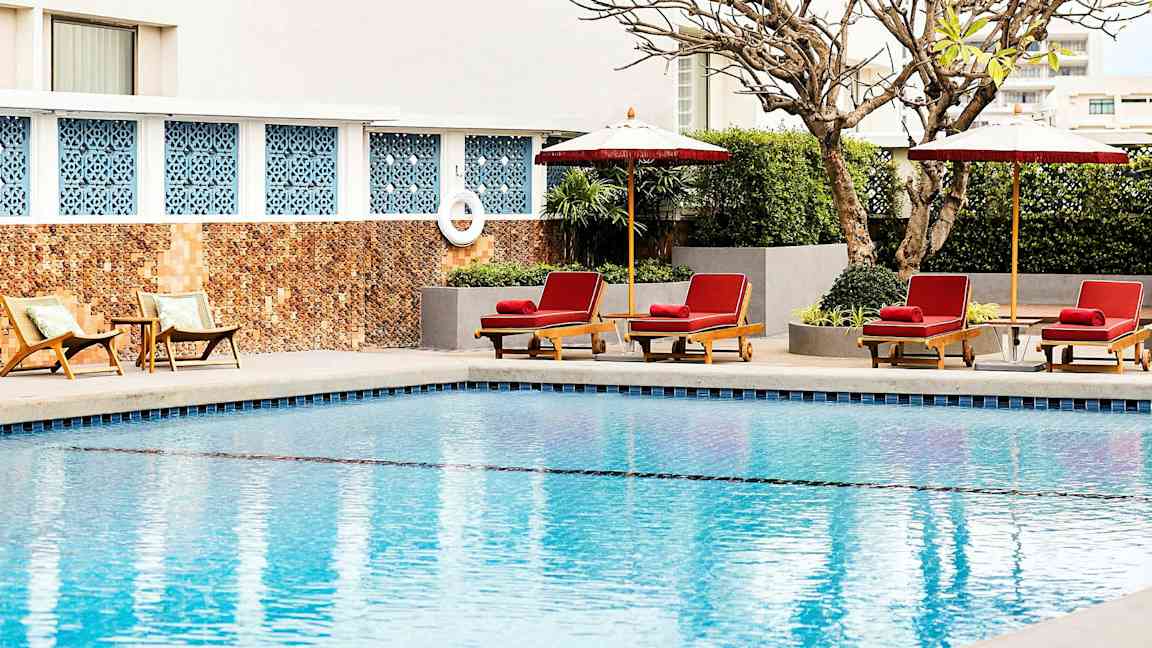 Et basseng med solsenger og parasoller ved kanten, omgitt av planter og en bygningsvegg i bakgrunnen. (AI-generert tekst)