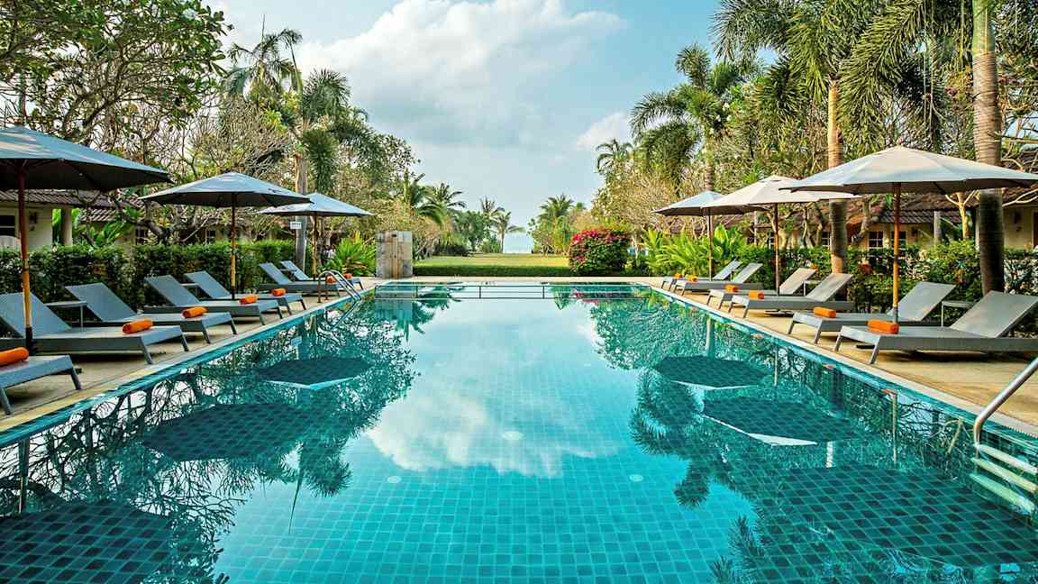 Bilde av et utendørsbasseng omgitt av solsenger, parasoller og grøntområder under en klar himmel. (AI-generert tekst)