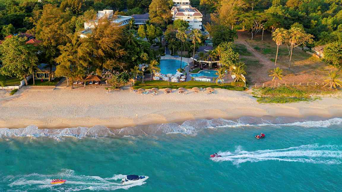 En strand med parasoller og stoler samt et hotellkompleks og bassenger i bakgrunnen omgitt av vegetasjon. (AI-generert tekst)