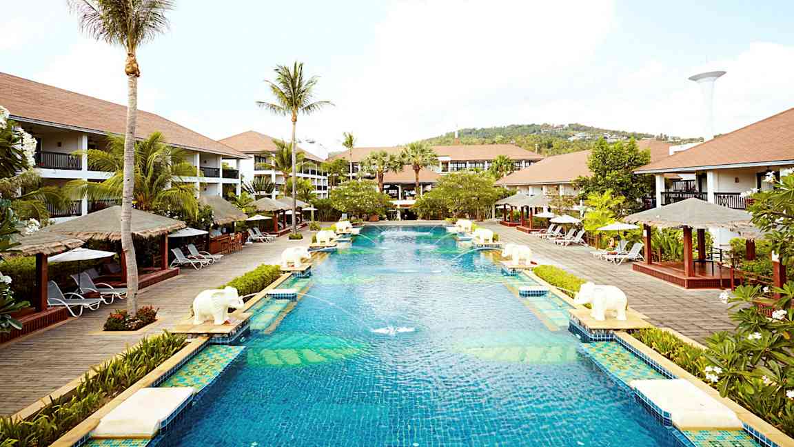 Et stort basseng på et feriested omgitt av palmer, solsenger og bungalowlignende bygninger. (AI-generert tekst)
