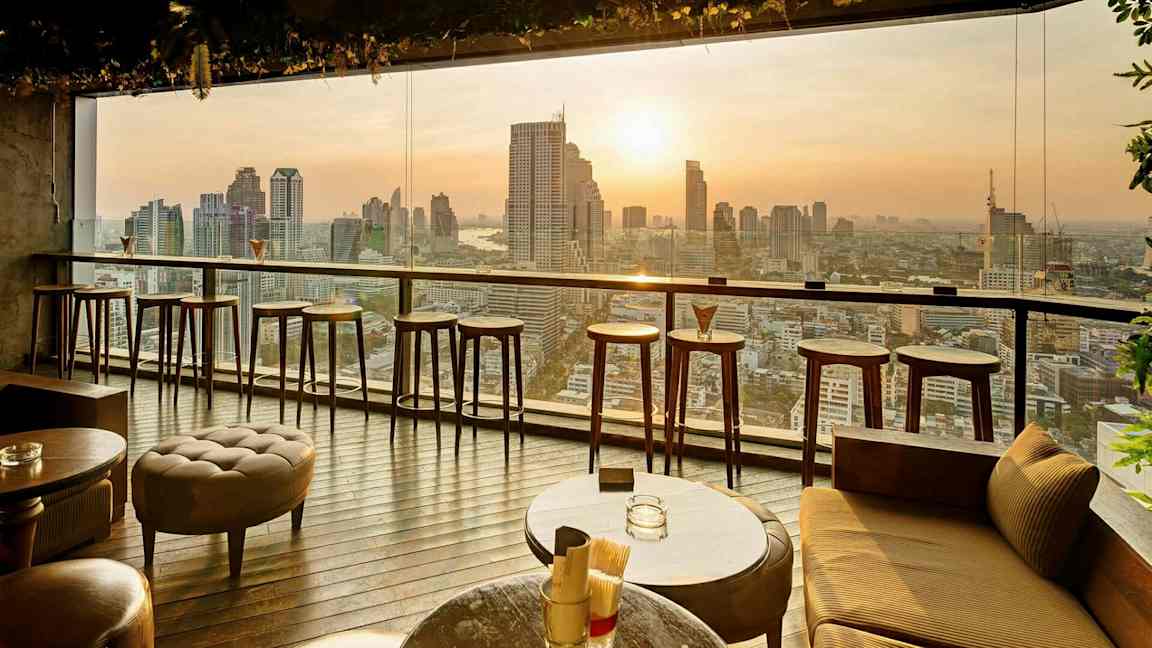 En moderne innendørs bar med høye barkrakker og bord foran et stort vindu med utsikt over en bysilhuett ved solnedgang. (AI-generert tekst)