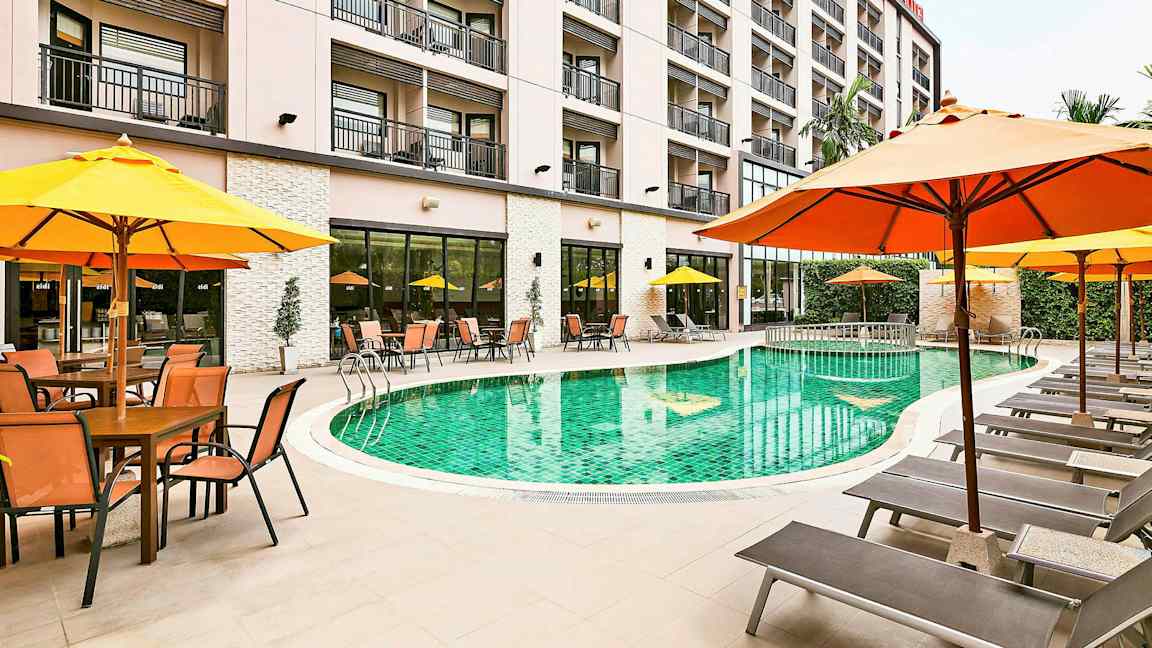 Et hotellbasseng omgitt av solsenger og parasoller, med bygningen i bakgrunnen. (AI-generert tekst)