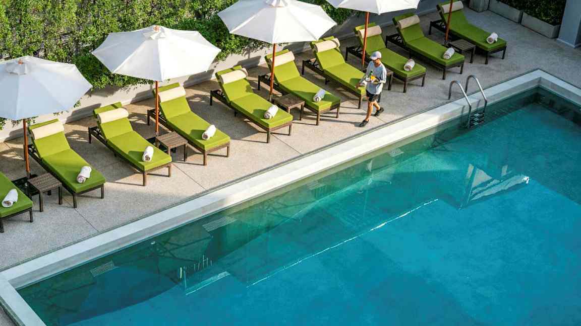 Basseng med solsenger og parasoller ved kanten, samt en person som går forbi. (AI-generert tekst)
