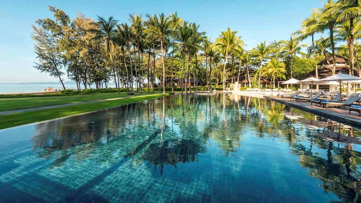 Et utendørsbasseng omgitt av grøntområder og palmer, med utsikt over stranden og havet. (AI-generert tekst)