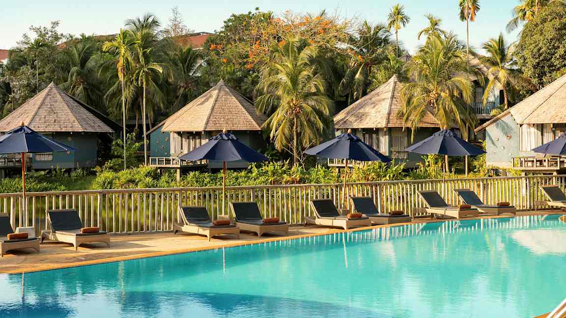 Solstoler og parasoller ved et svømmebasseng med bungalowlignende bygninger og vegetation i bakgrunnen. (AI-generert tekst)