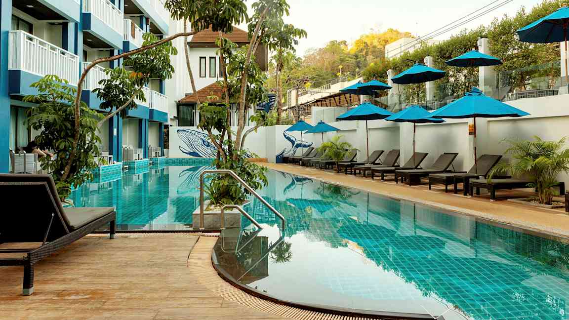 Et utendørs svømmebasseng med solsenger og parasoller, omgitt av vegetasjon og en hotellbygning med flere etasjer. (AI-generert tekst)