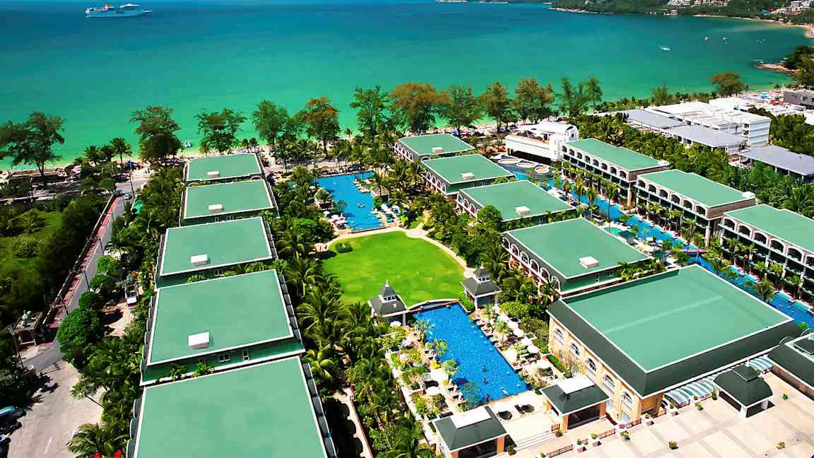 Oversiktsbilde av et feriehotell med flere bygninger omgitt av grøntområder og bassenger, beliggende ved havet. (AI-generert tekst)