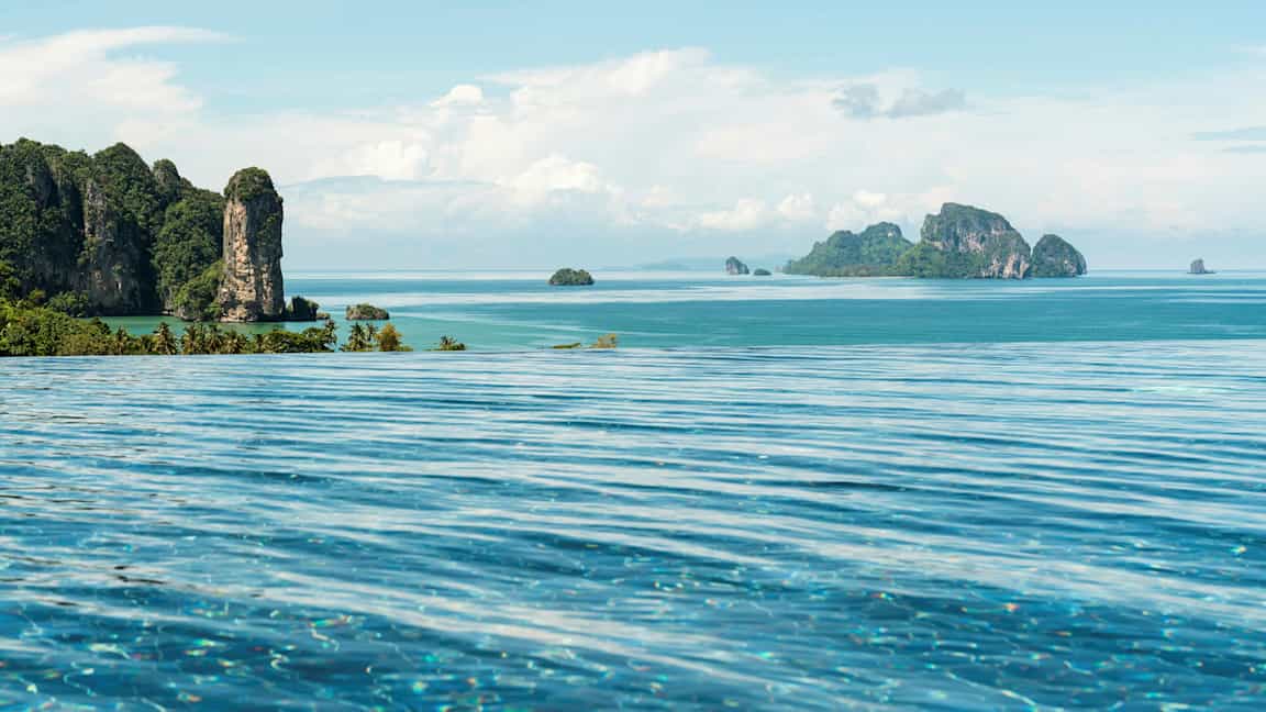 Et evighetsbasseng i forgrunnen med utsikt over en tropisk kyst- og havutsikt med fjellrike øyer. (AI-generert tekst)