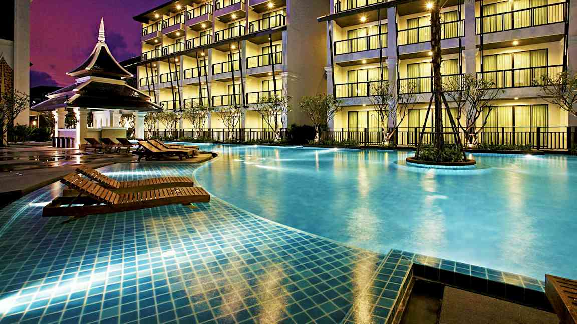 En opplyst hotellfasade med balkonger i flere etasjer, foran et basseng med tresolsenger og en liten paviljong. (AI-generert tekst)