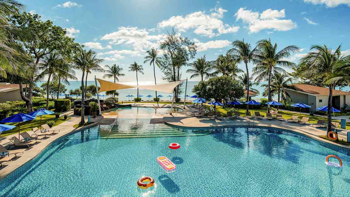 Et svømmebasseng omgitt av solsenger og parasoller, med utsikt over havet og palmer under en klar himmel. (AI-generert tekst)