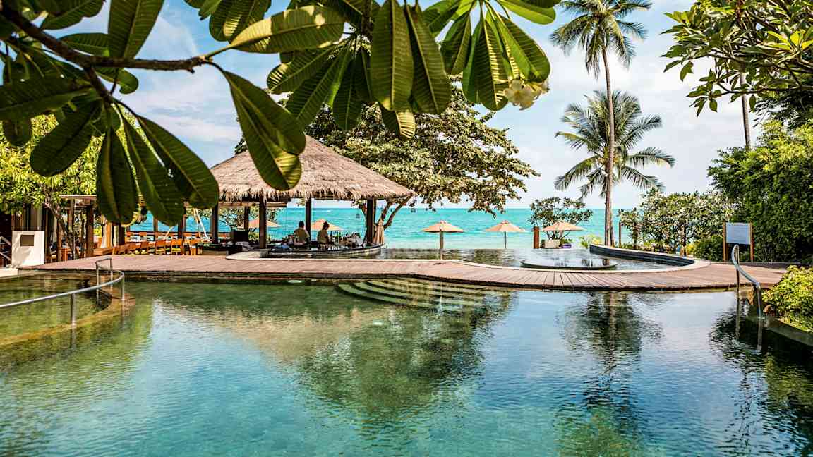 Stor basseng i et tropisk miljø med trær, palmer og havet i bakgrunnen. (AI-generert tekst)