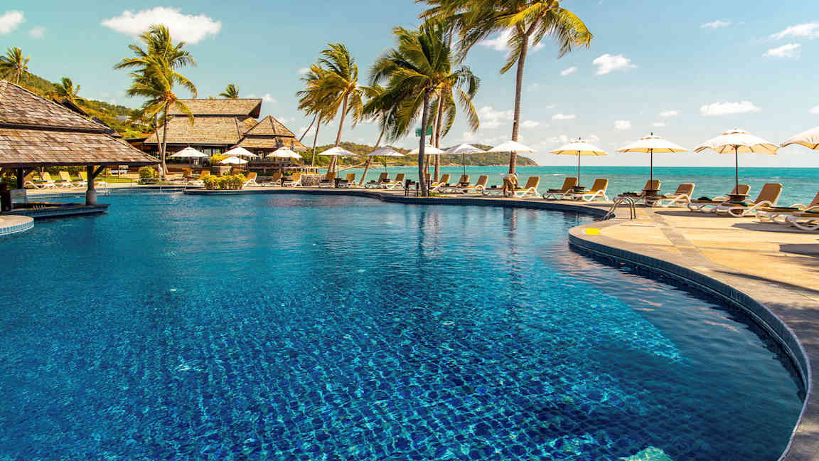 Bilde av et utendørsbasseng omgitt av solsenger og palmer, med havet i bakgrunnen. (AI-generert tekst)