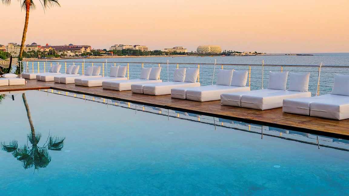Solstoler ved et basseng med utsikt over havet og bygninger i bakgrunnen. (AI-generert tekst)