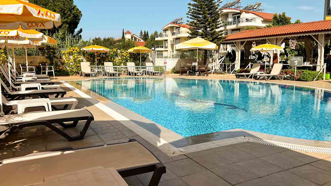 Et utendørs svømmebasseng med solsenger og parasoller, omgitt av grøntområder og bygninger. (AI-generert tekst)