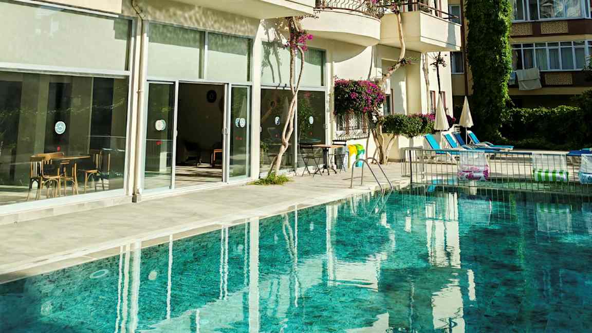 Et basseng foran en bygning med balkonger og store glassvinduer. Bildene viser også stoler og bord samt parasoller ved bassengområdet. (AI-generert tekst)