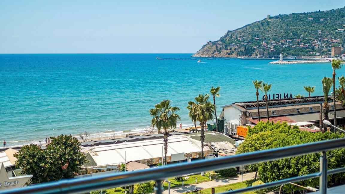 Utsikt over havet og en strand med bygninger i forgrunnen. En odde med bebyggelse er synlig i bakgrunnen. (AI-generert tekst)