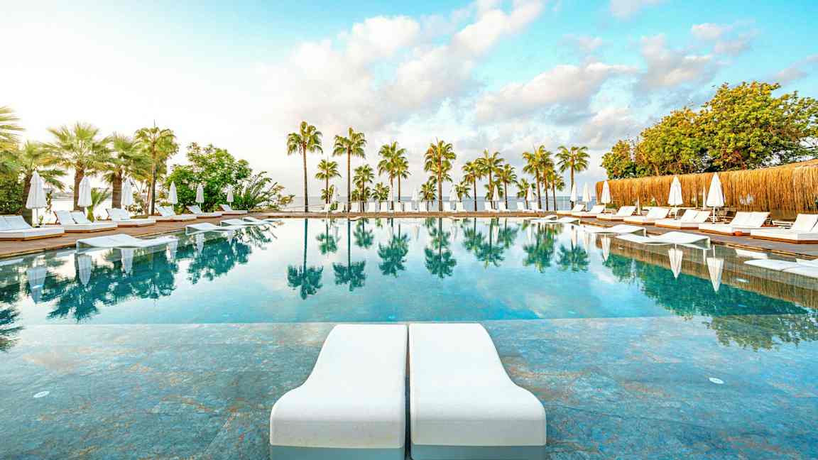 Et utendørsbasseng med solsenger og parasoller, omgitt av palmer, og med speilinger i vannet. (AI-generert tekst)