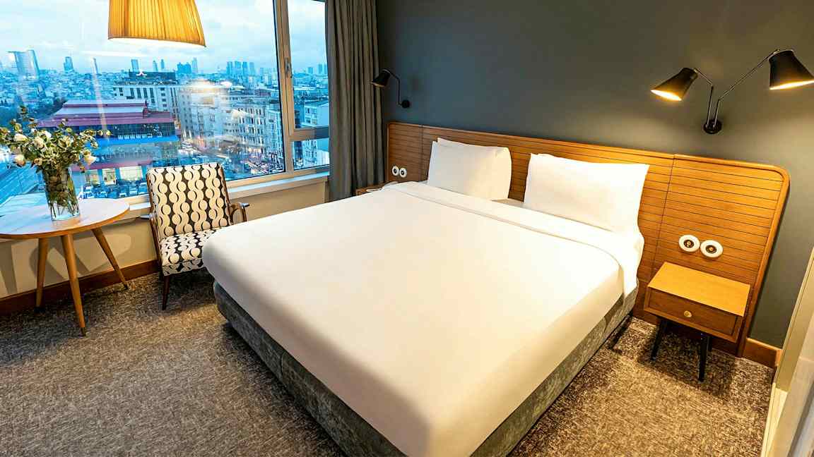 Et hotellrom med en stor seng, vindu og utsikt over en by. (AI-generert tekst)