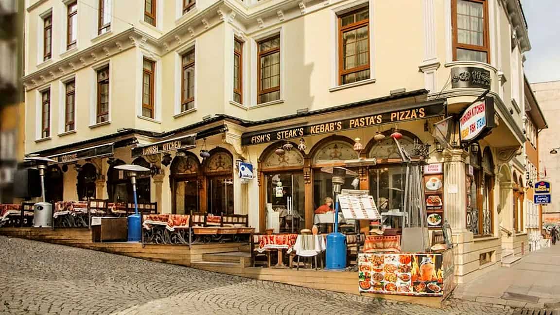 En gate med en bygning som har restaurant med uteservering, skilt med mat som kebab og pizza. (AI-generert tekst)