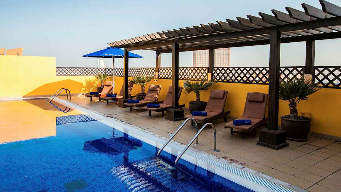 Et utendørsbasseng med blått vann omgitt av solsenger og parasoller. Solsengene står på et belagt gulv med en gul vegg og trepergola i bakgrunnen. (AI-generert tekst)