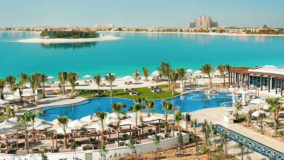 Et luksuriøst feriested med et basseng og strand ved en blå lagune, med en ø i bakgrunnen. (AI-generert tekst)