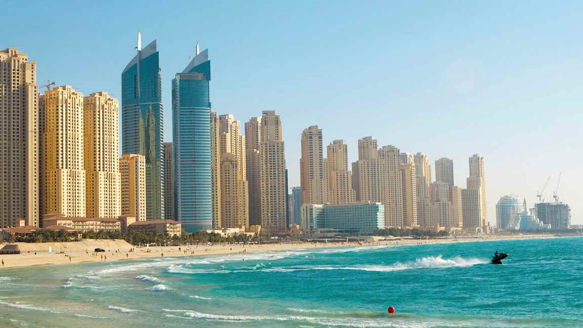 Bilde av høyhus ved en strand med klart blått vann, en person er på en jetski nær stranden. (AI-generert tekst)