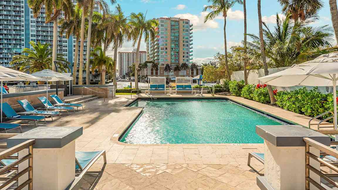 Et svømmebasseng omgitt av solsenger og parasoller, med høye bygninger og palmer i bakgrunnen. (AI-generert tekst)