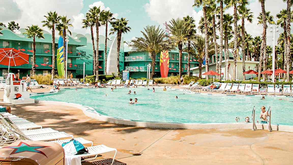 Et utendørs basseng omgitt av solsenger, parasoller og palmer. I bakgrunnen sees en bygning med fargerike detaljer. (AI-generert tekst)