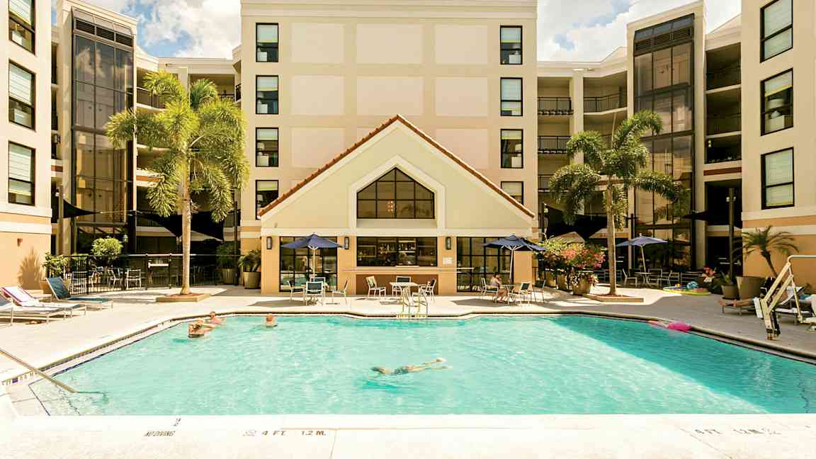 Bilde av et hotell med et basseng i forgrunnen. Personer svømmer og slapper av ved bassengområdet. Hotellfasaden er synlig i bakgrunnen med palmer i nærheten. (AI-generert tekst)