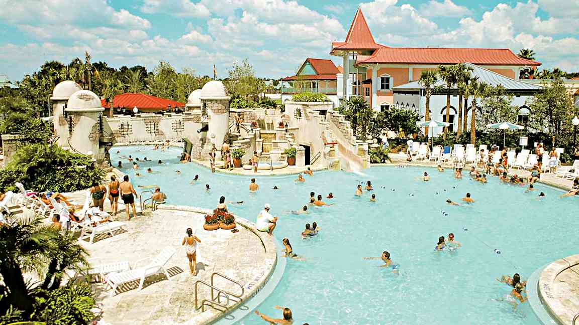 Et utendørsbasseng omgitt av mennesker, solsenger og bygninger. (AI-generert tekst)