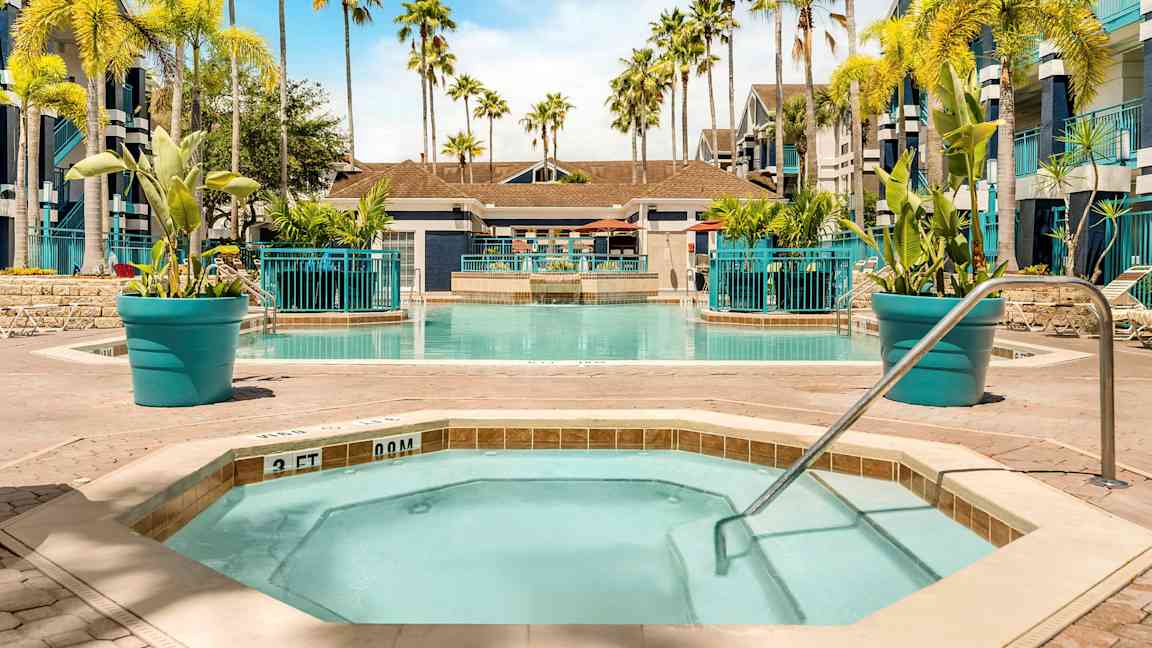 Bilde av et basseng omgitt av solsenger og palmer. I forgrunnen er det et mindre boblebad med gelender. (AI-generert tekst)