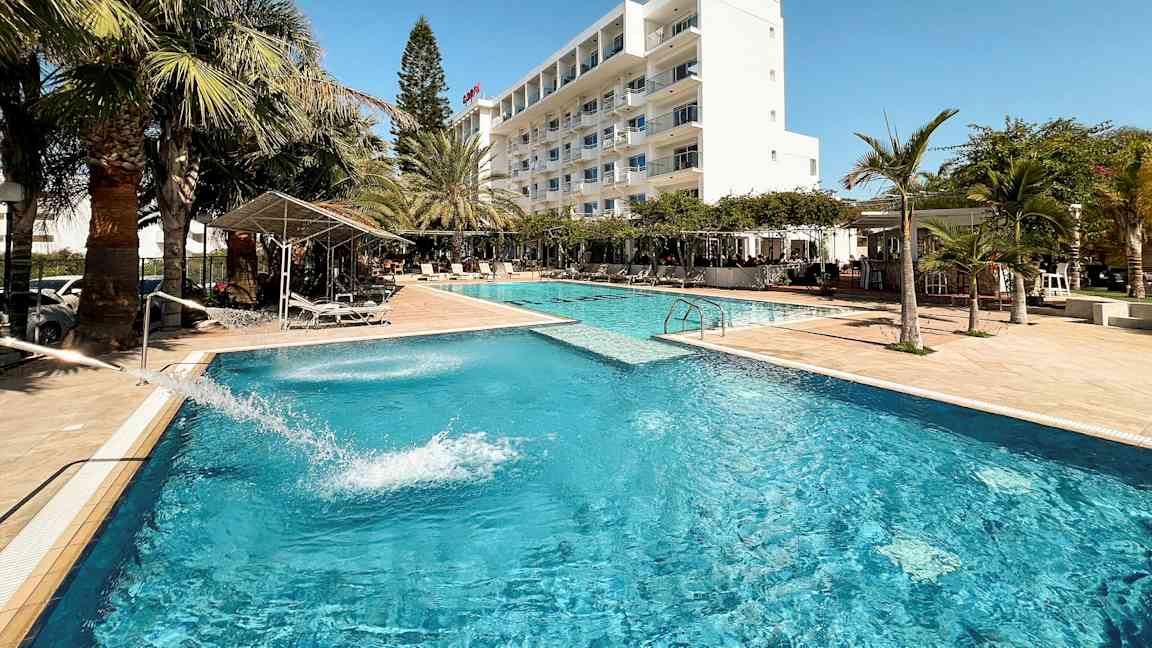 Et hotellbasseng omgitt av palmer og en hvit bygning i bakgrunnen. (AI-generert tekst)