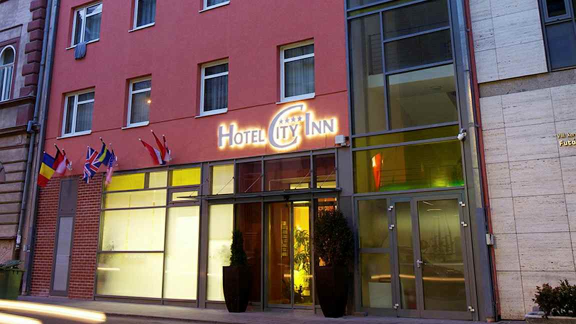 Utenfor en hotellbygning med et skilt som lyder "Hotel City Inn" og flere flagg ved inngangen. (AI-generert tekst)