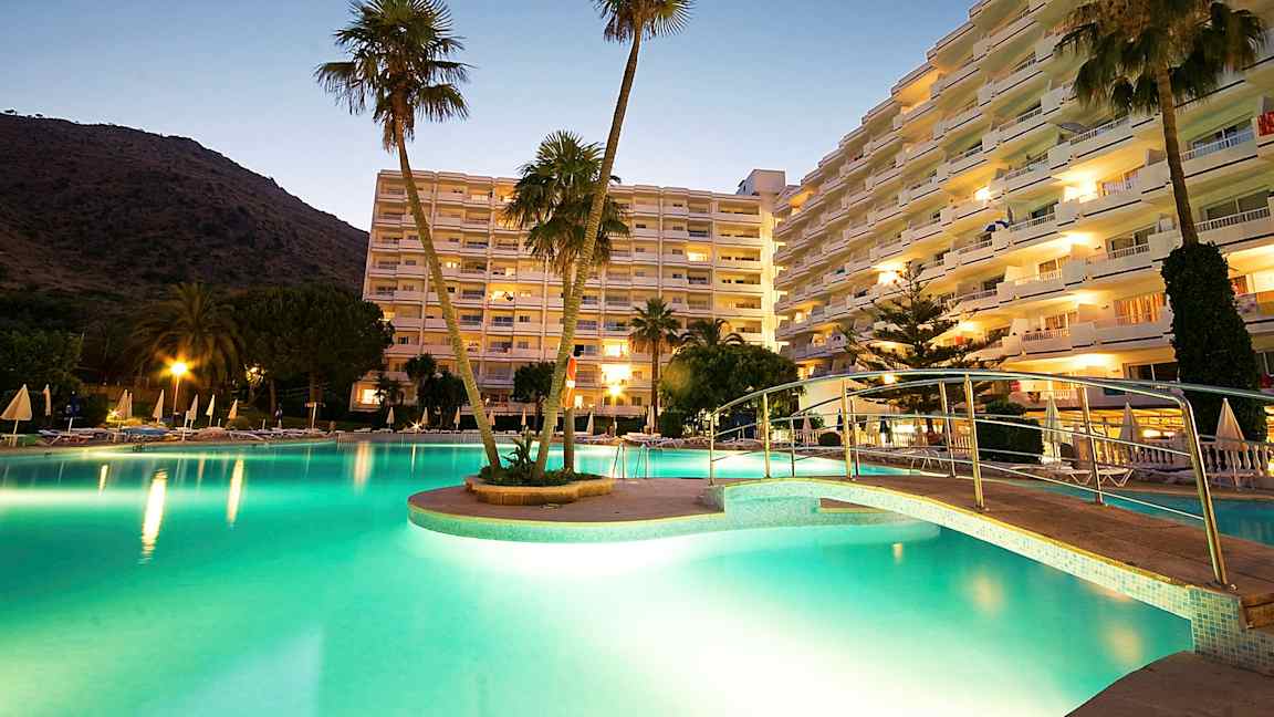 Et bilde av et hotellområde ved skumring, med et opplyst basseng, en bro og høye bygninger i bakgrunnen. (AI-generert tekst)