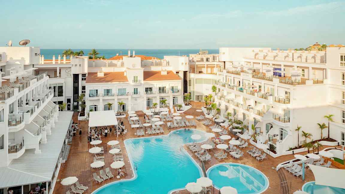 Utsikt over hotellområde med basseng og solsenger, havet er synlig i bakgrunnen. (AI-generert tekst)