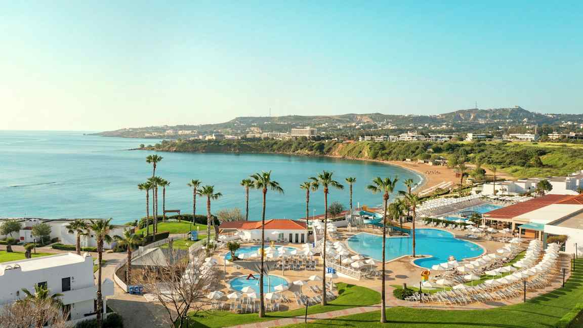 Utsikt over hotellområde med bassenger og utsikt mot en bukt med blått vann og grønne omgivelser. (AI-generert tekst)