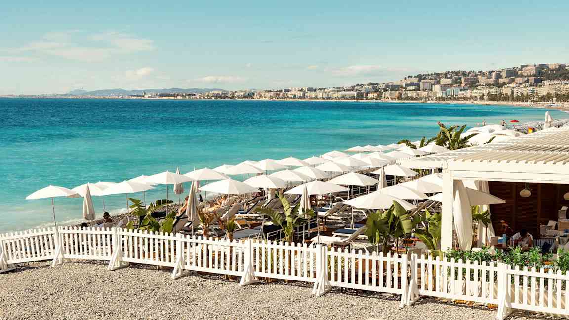 En solrik strand med blått hav, parasoller og sitteplasser omgitt av et hvitt gjerde. (AI-generert tekst)
